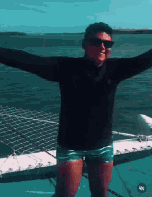 a man wearing sunglasses and a black shirt stands on a boat with his arms outstretched