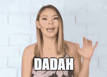 a woman in a floral dress is waving her hand in front of a brick wall with the word dadah written on it .