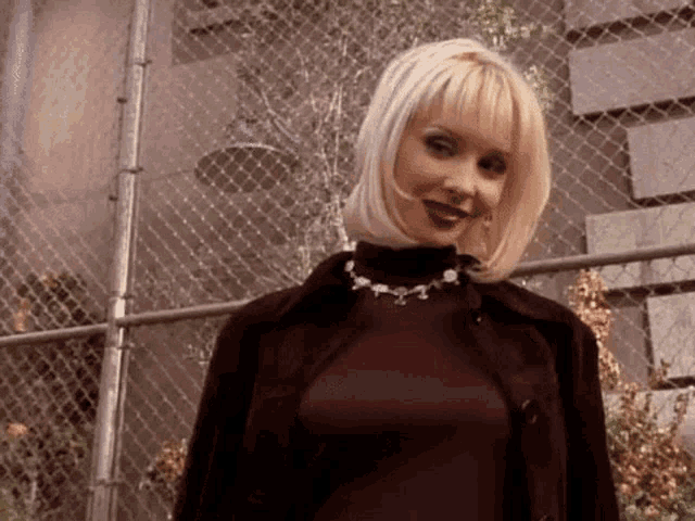 a woman with blonde hair and a necklace is standing in front of a fence