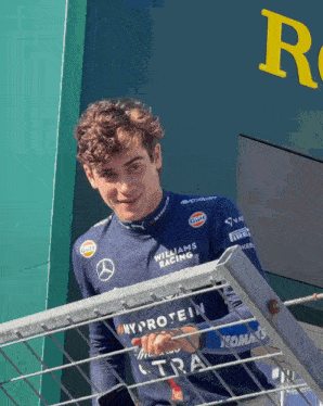 a young man wearing a williams racing shirt is leaning on a railing