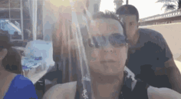 a man wearing sunglasses looks at the camera while another man stands behind him