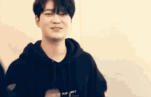 a young man in a black hoodie is smiling while standing in front of a wall .