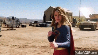 a woman in a superman costume is standing in the middle of a dirt field holding a dollar bill .