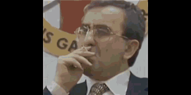 a man in a suit and tie is smoking a cigarette in front of a sign that says ' s ga '