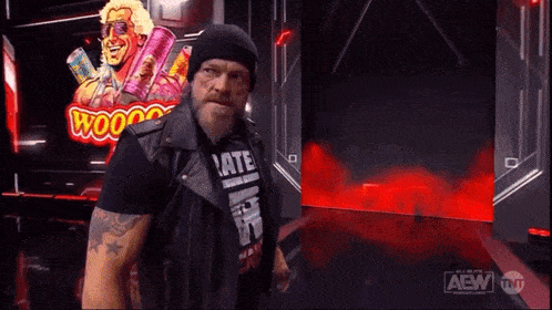 a man in a leather vest and beanie is standing in front of a sign that says aew .