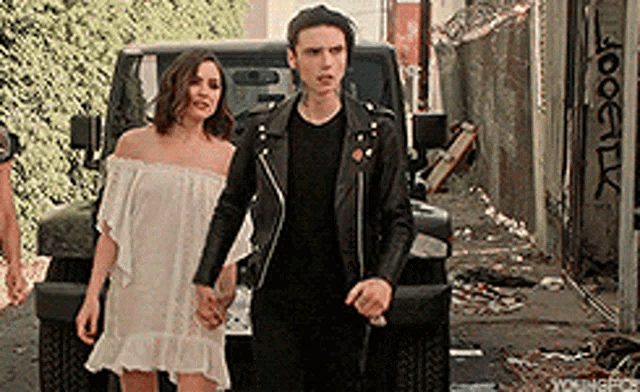 a man and a woman are holding hands in front of a jeep . the woman is wearing a white dress .
