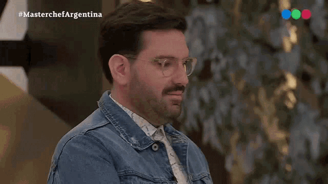 a man with glasses and a beard is wearing a denim jacket and a shirt .