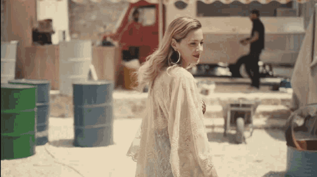 a woman wearing hoop earrings stands in front of barrels on a beach