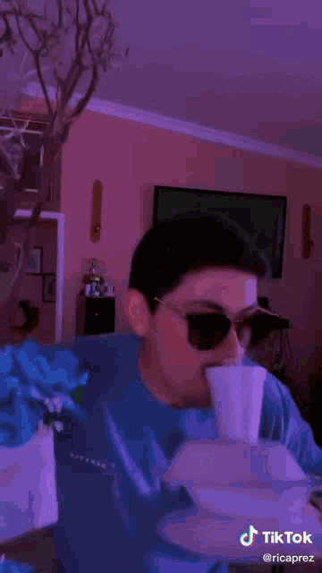 a man wearing sunglasses and a blue shirt is drinking from a styrofoam cup ..