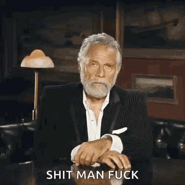 a man in a suit is sitting at a table and covering his face with his hand with the words shit man fuck written in front of him