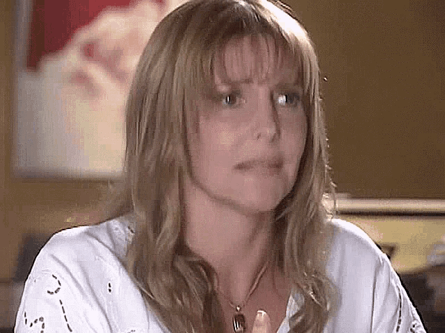 a woman with long blonde hair is wearing a white shirt and a necklace and making a funny face .