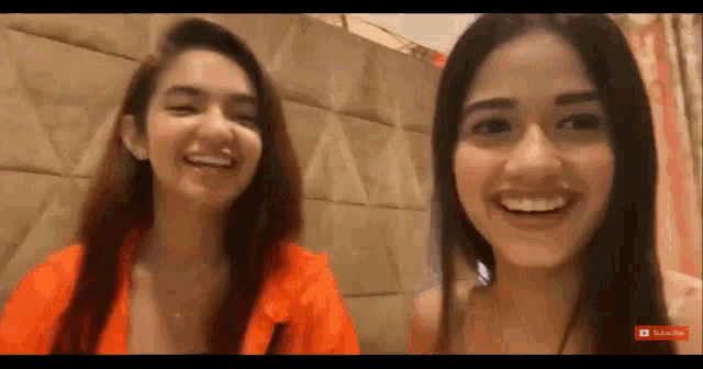 two women are smiling and laughing in front of a wall . one of the women is wearing an orange shirt .