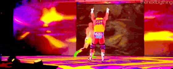 a wrestler is standing on a stage with his arms in the air while another wrestler is standing behind him .