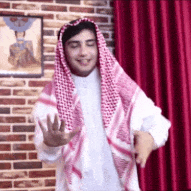 a man wearing a keffiyeh and a white shirt is standing in front of a brick wall and red curtain .