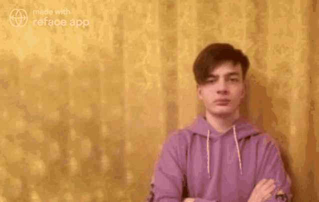 a young man in a purple hoodie is standing in front of a yellow curtain with his arms crossed .