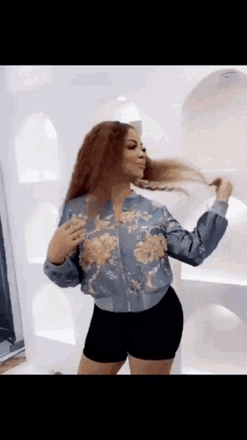 a woman in a blue jacket and black shorts is standing in front of a white wall holding her hair .