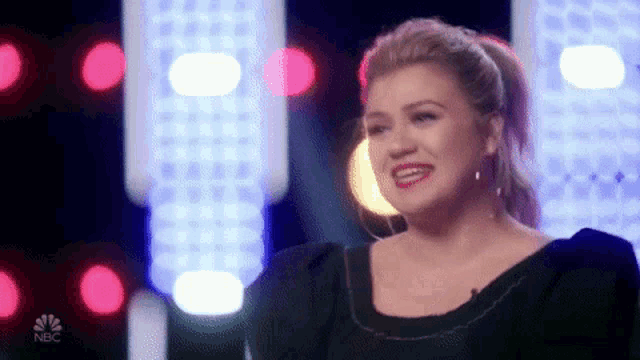 a woman in a black dress is smiling while sitting on a stage in front of a crowd .