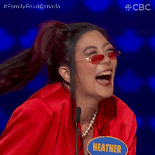 a woman wearing sunglasses and a red jacket is laughing with a blue sign that says heather