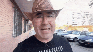 a man wearing a cowboy hat and glasses is standing in front of a brick building and a parking lot .