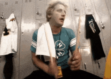 a man sitting in a locker room holding a drum stick and a towel around his neck with the letter h on it