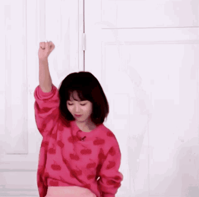 a woman in a pink sweater is standing in front of a white wall with her fist in the air .