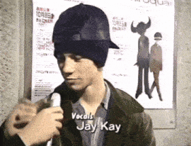 a man wearing a hat is talking into a microphone with the name jay kay on the bottom right