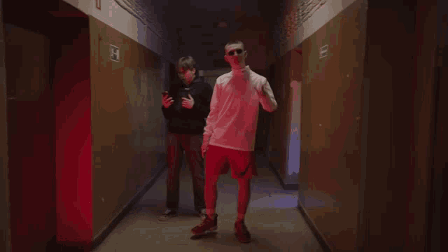 two men are standing in a hallway with a red exit sign on the wall