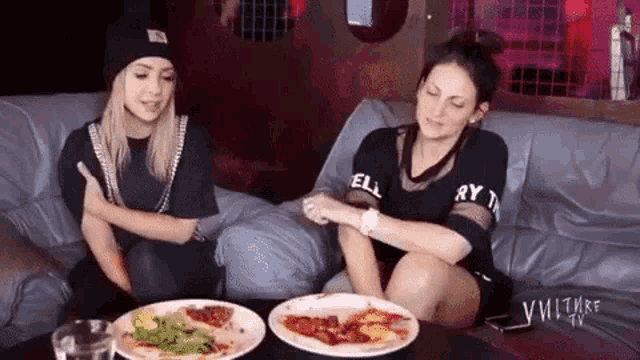 two women are sitting on a couch eating food .