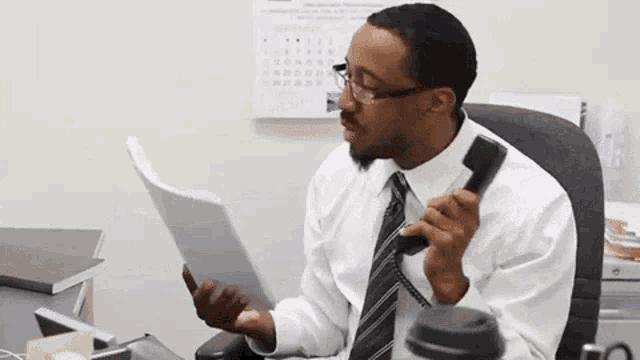 a man sitting at a desk talking on a phone while holding a piece of paper