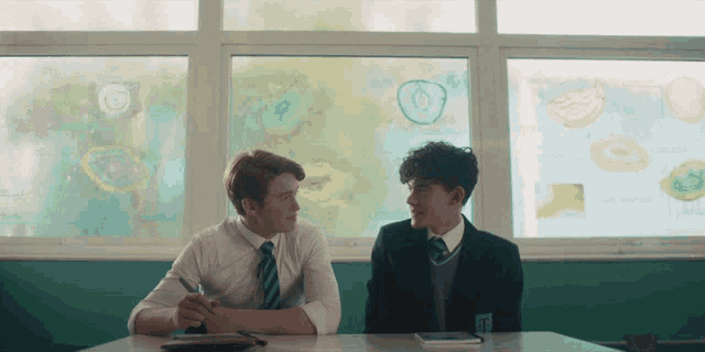 two boys are sitting at a desk in front of a window with drawings on the window