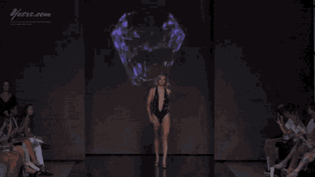 a woman walks down a runway wearing a very plunging black swimsuit