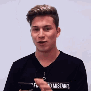 a young man in a black t-shirt is holding a cell phone and making a funny face .