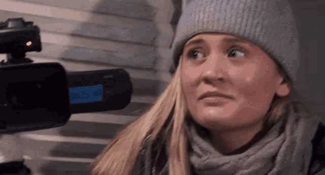 a woman wearing a hat and scarf is looking at a video camera .