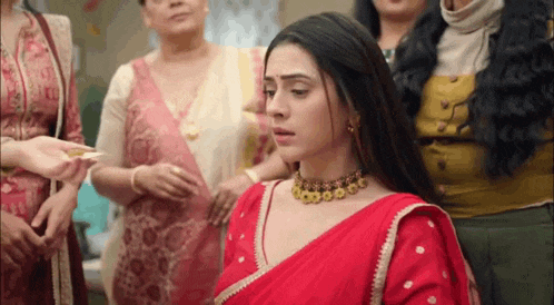 a woman in a red saree and a gold necklace is standing in a room with other women .