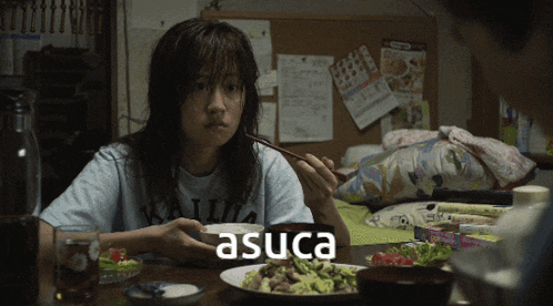a woman sitting at a table with chopsticks and a plate of food with the word asuca on it