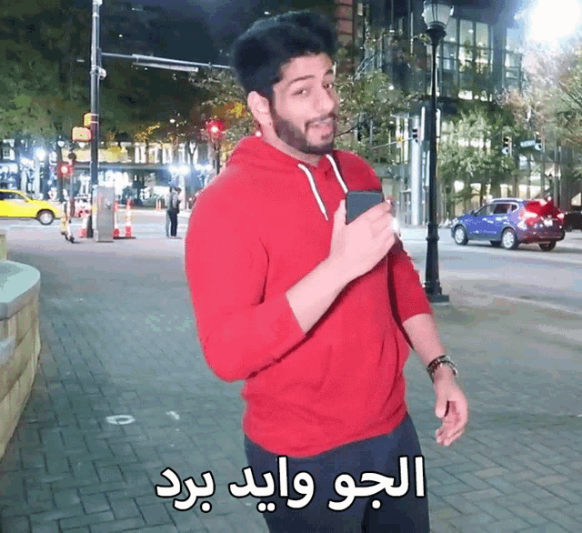 a man in a red hoodie is standing on a sidewalk holding a cell phone with arabic writing on it