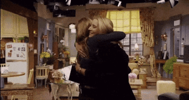two women hugging each other in a living room with a refrigerator in the background