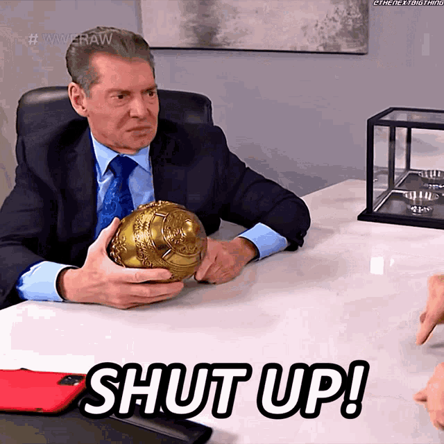 a man in a suit and tie is sitting at a table holding a gold object and the words shut up are above him
