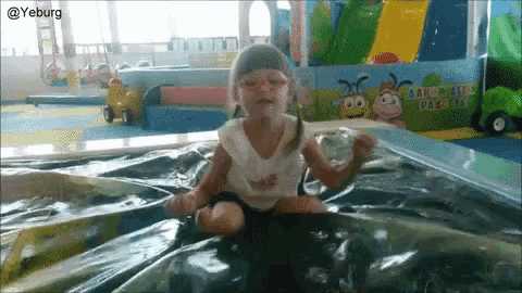 a little girl is sitting on a plastic sheet in a playground with a cartoon character on the wall behind her ..