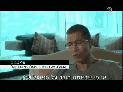 a man wearing glasses is sitting on a couch in front of a window with the number 2 on the screen