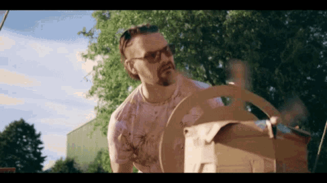 a man wearing sunglasses and a pink shirt holds a cardboard box