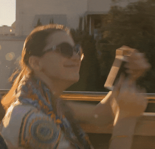 a woman wearing sunglasses and a scarf smiles while holding a camera