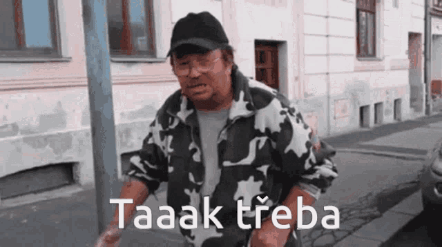 a man in a camouflage jacket is standing on a street with the words taaak třeba written on the sidewalk