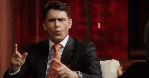a man in a suit and tie is sitting on a couch making a funny face and pointing at the camera .