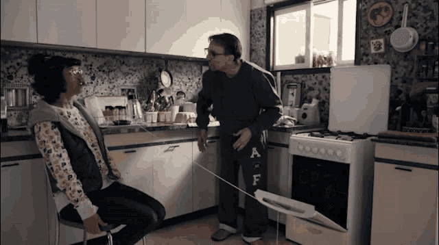a man standing next to a woman in a kitchen wearing a pair of pants with the letter a on them