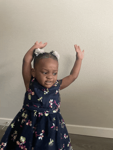 a little girl in a blue dress with flowers on it stands with her hands in the air