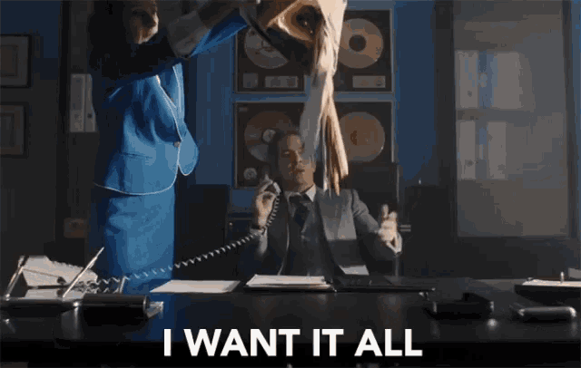 a man sitting at a desk talking on a phone with the words i want it all written on the screen behind him