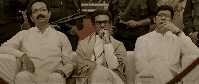 three men are sitting next to each other with one wearing glasses and a necklace
