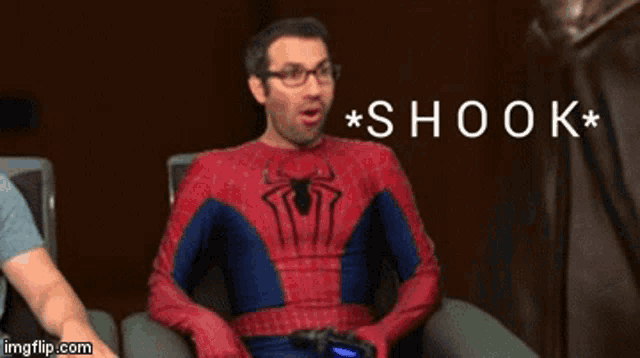 a man in a spiderman costume is holding a controller and says shook