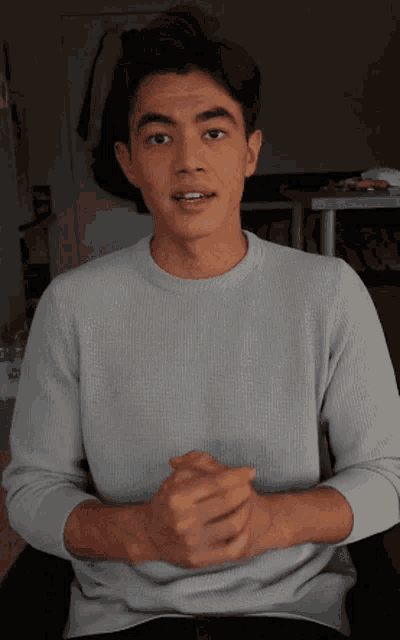 a young man in a grey sweater is sitting with his hands folded